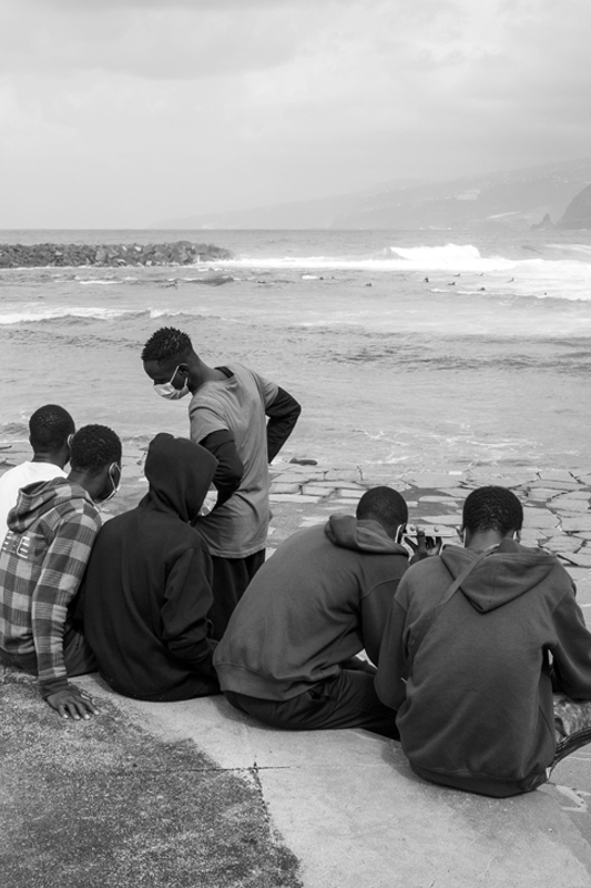 Grupa uchodźców siedzi na falochronie, w tle wzburzone morze.