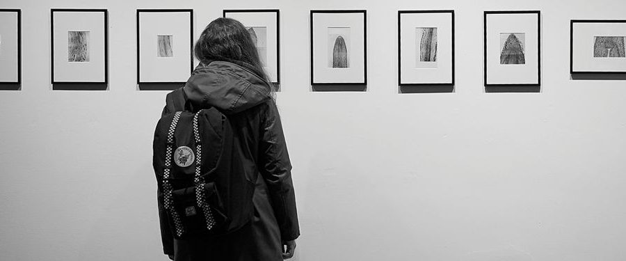 Osoba stojąca tyłem do obiektywu, przed obrazkami zawieszonymi na ścianie. Zdjęcie czarno-białe.