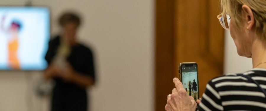 Zdjęcie przedstawia wnętrze Galerii Promocyjnej a w niej dwie osoby: na pierwszym planie kobieta w okularach robi zdjęcie telefonem innej osobie - postać rozmyta- która stoi obok ekranu wyświetlającego instalację video będącą częscią wystawy.