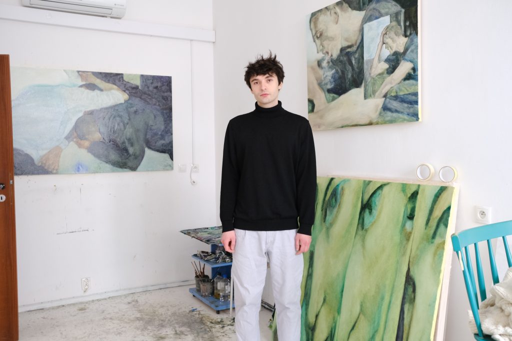 autor wystawy, Bartosz Kowal w białych spodniach i czarnej bluzie stoi w swojej pracowni, w tle obrazy, z prawej niebieskie krzesło